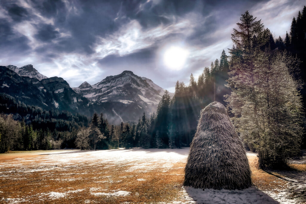 paysage d'hiver avec un peu de neige au sol et le soleil qui irradie derrière les nuages sur fond de montagne avec une grosse botte de foin